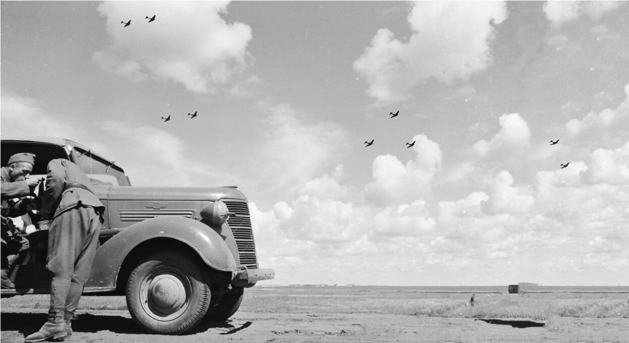 Truck and two soldiers by the roadside in 1941 with bombers flying overhead