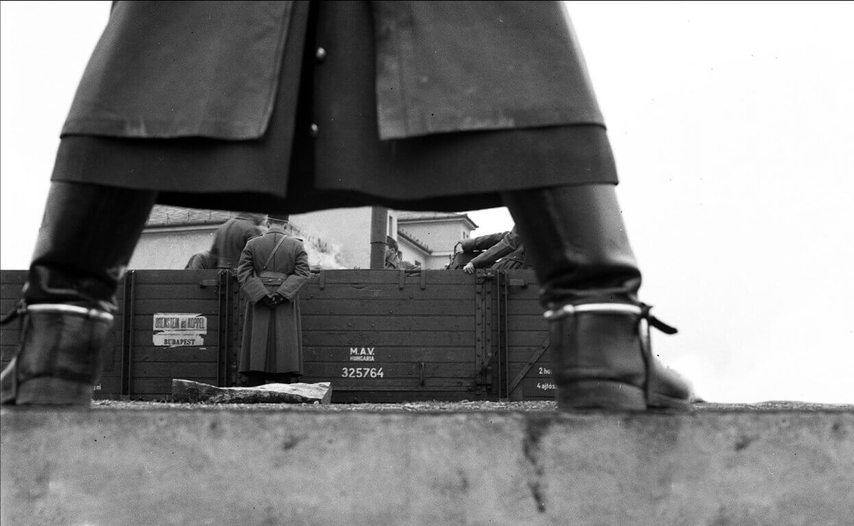 An intimidating Hungarian soldier leather boots and trench coat straddling a stone wall in 1941