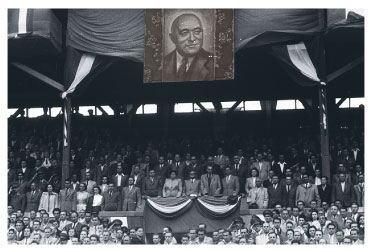 communist leadership watching football under a large portrait of Mátyás Rákosi